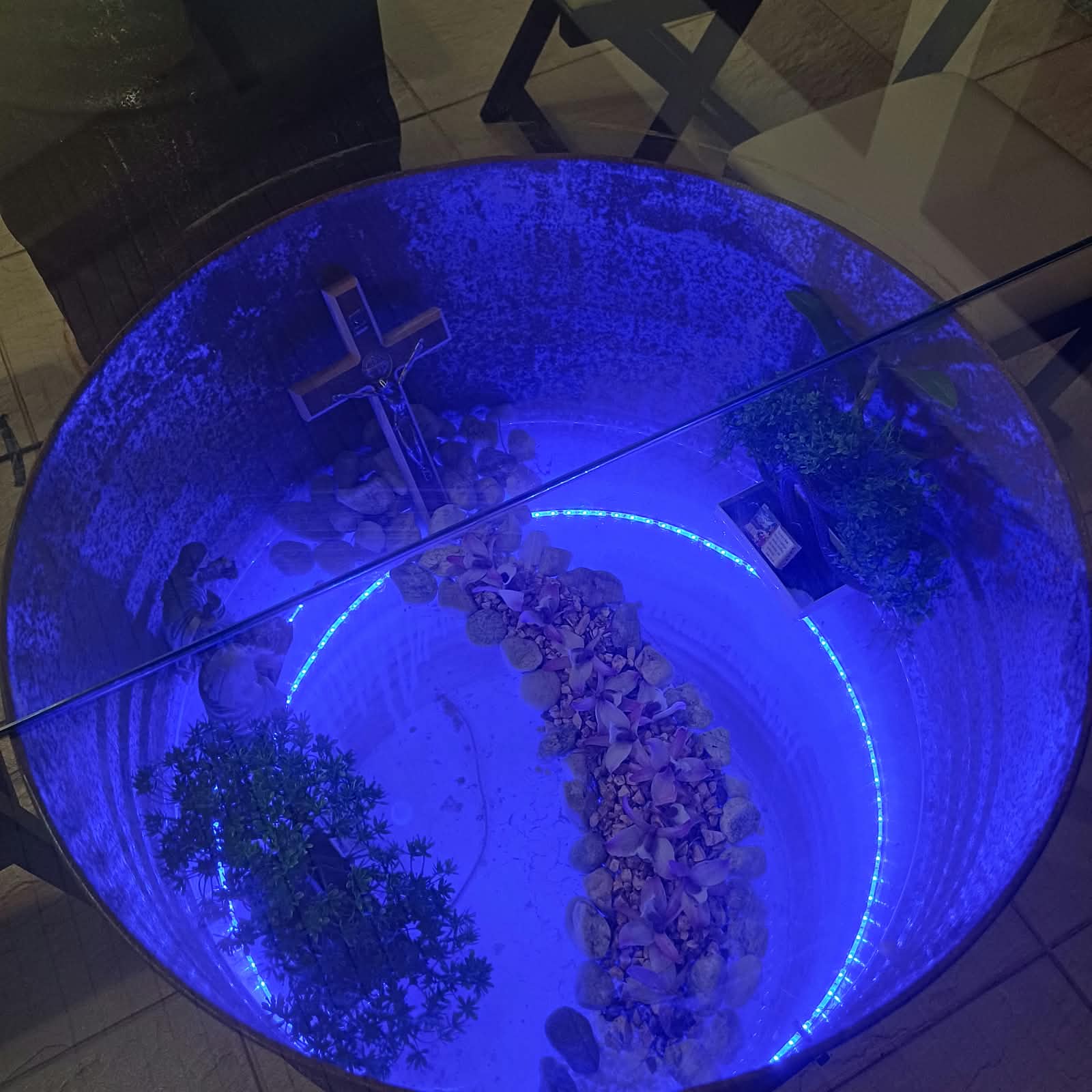 Detalhe da Mesa Kairós: cruz de madeira iluminada em azul, cercada por pedras e flores, dentro do tampo da mesa